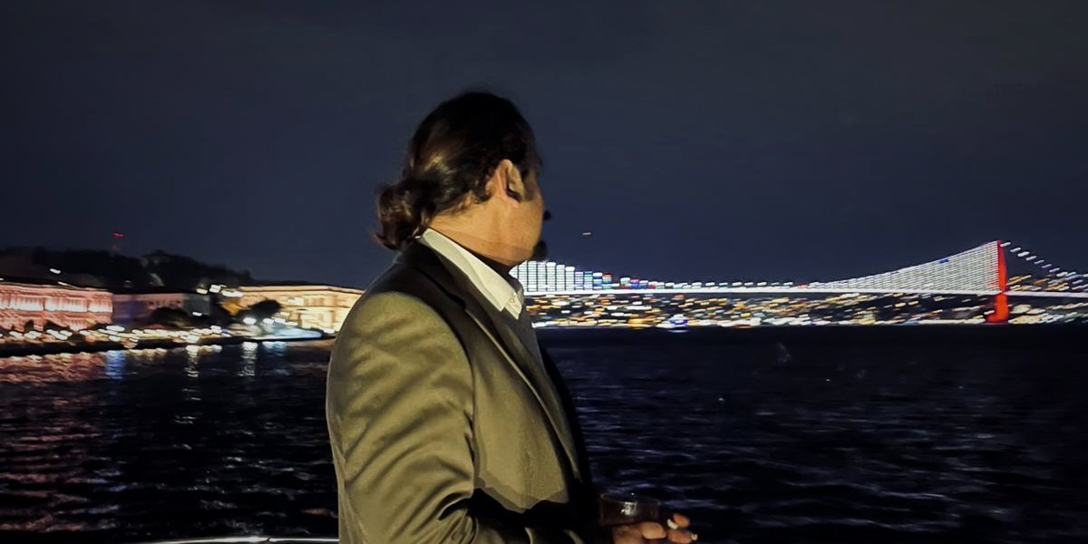 Doğan Özhan on a yacht deck at night, looking toward the lit Bosphorus Bridge